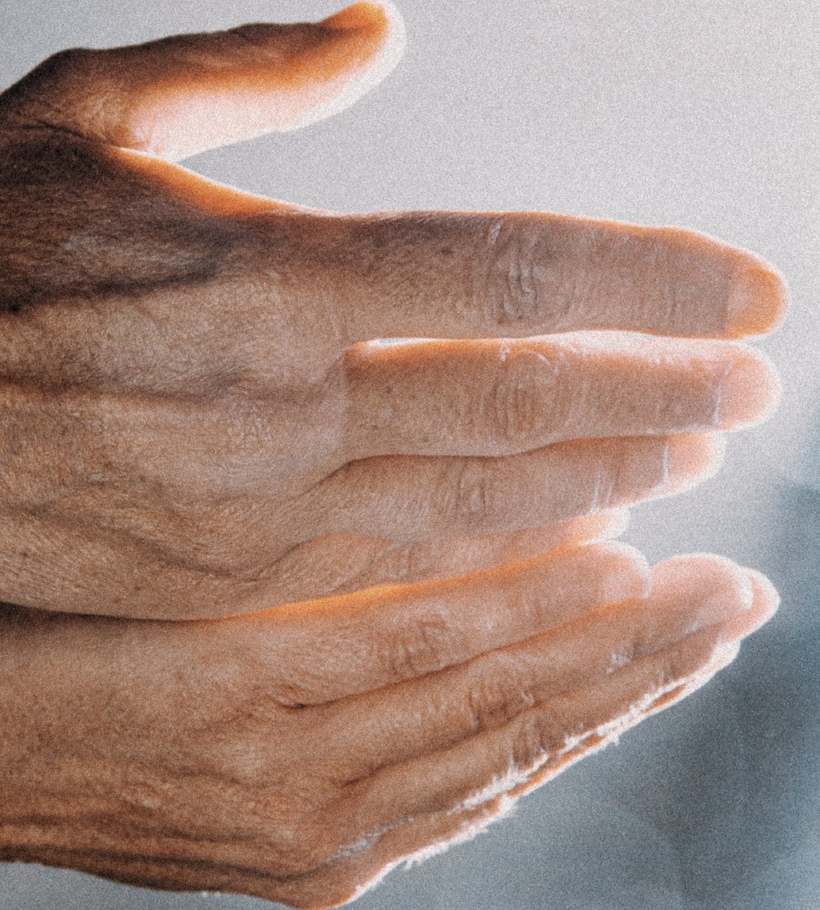 Duas mãos abertas, de pele marcada pelo sol, estendidas na luz sobre um fundo cinza-azulado